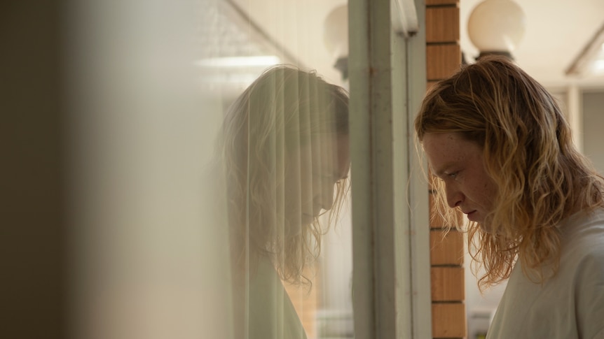 A young man with shoulder-length blonde hair looks despondently at his reflection in the window of a brick house.