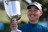 Min Woo Lee smiles while holding a large silver trophy