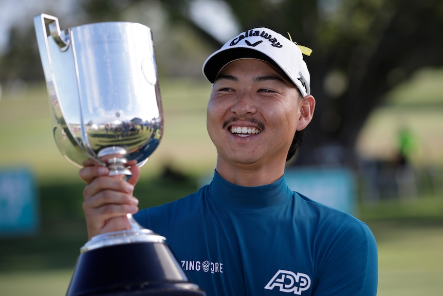 Min Woo Lee smiles while holding a large silver trophy