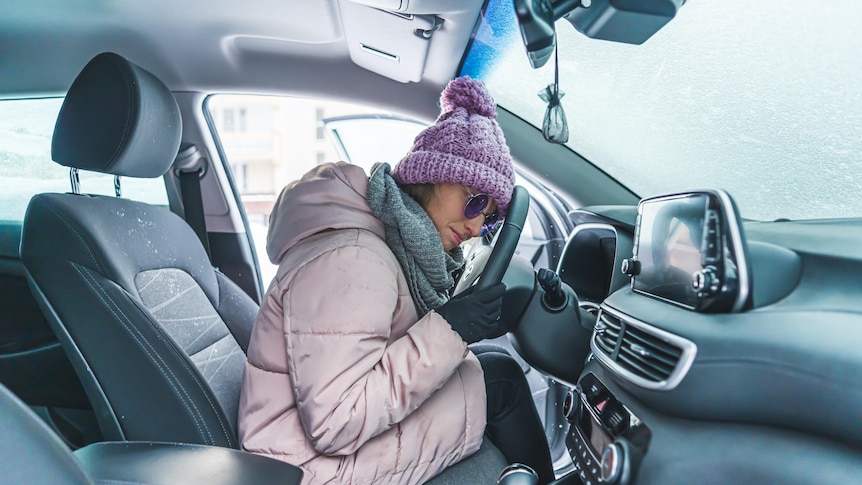 A woman wearing a beanie, puffer jacket, scarf and gloves sits in her car, unable to move as the windscreen is frozen.