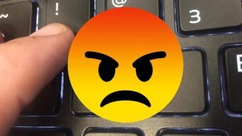 An angry-face emoji superimposed over a close up image of a finger typing an exclamation mark on a keyboard.