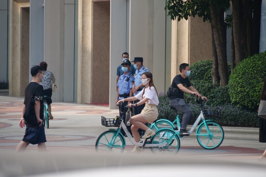 A woman rides her bike past a group of policemen standing guard near a building