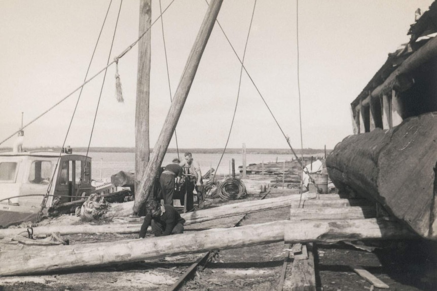 Historic image of men moving logs outside sawmill on waterfront