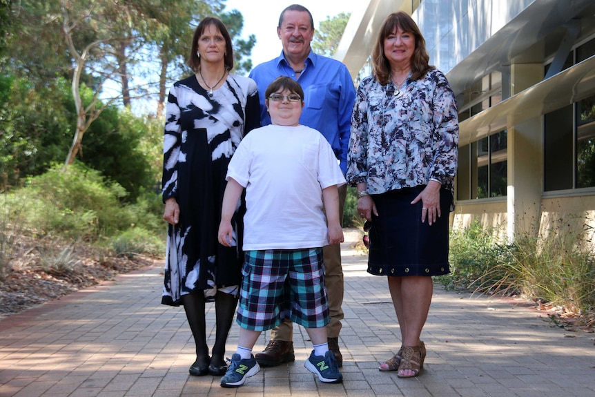 Billy stands with professors Sue Fletcher and Seve Wilton, and his mum, on a footpath, in front of some trees.
