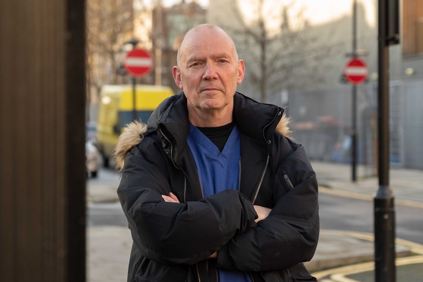 A middle-aged man stares into the camera with his arms crossed on a street in London.