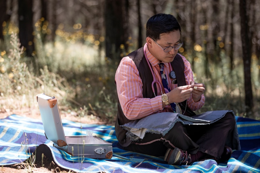 A man with short black hair and glasses sits on a blue picnic rug surrounded by bushland and sews by hand.