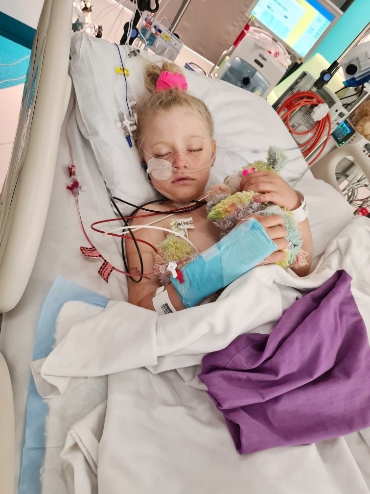 A young girl lying in a hospital bed cuddling a teddy bear with lots of cords and surgical tape on her