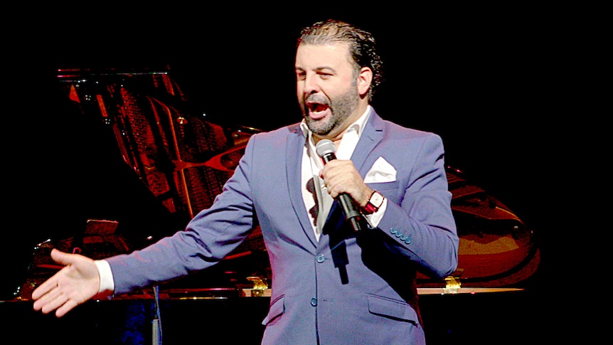 Opera singer David Serero performs on stage in a blue suit singing into a microphone.
