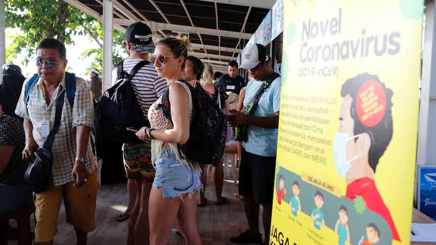 A group of people in sun glasses wait in a shaded area outside a building next to a yellow banner.