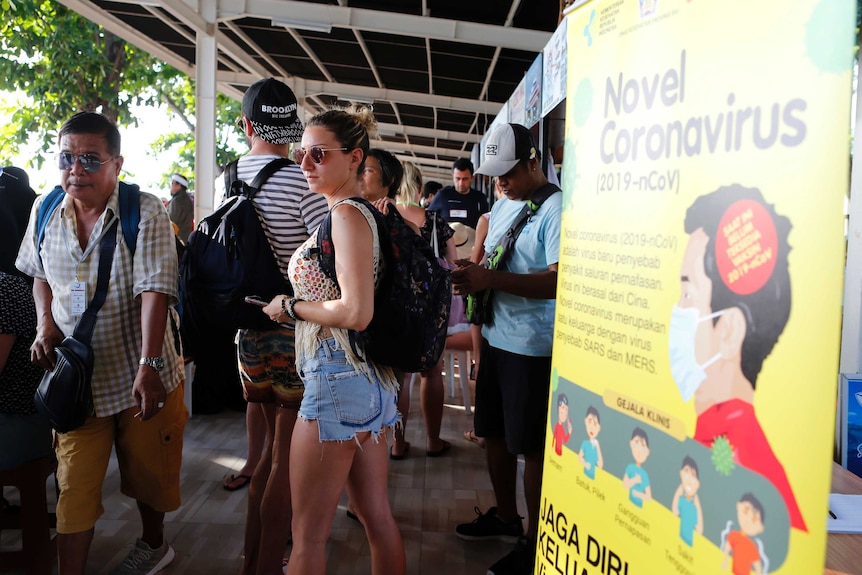 A group of people in sun glasses wait in a shaded area outside a building next to a yellow banner.