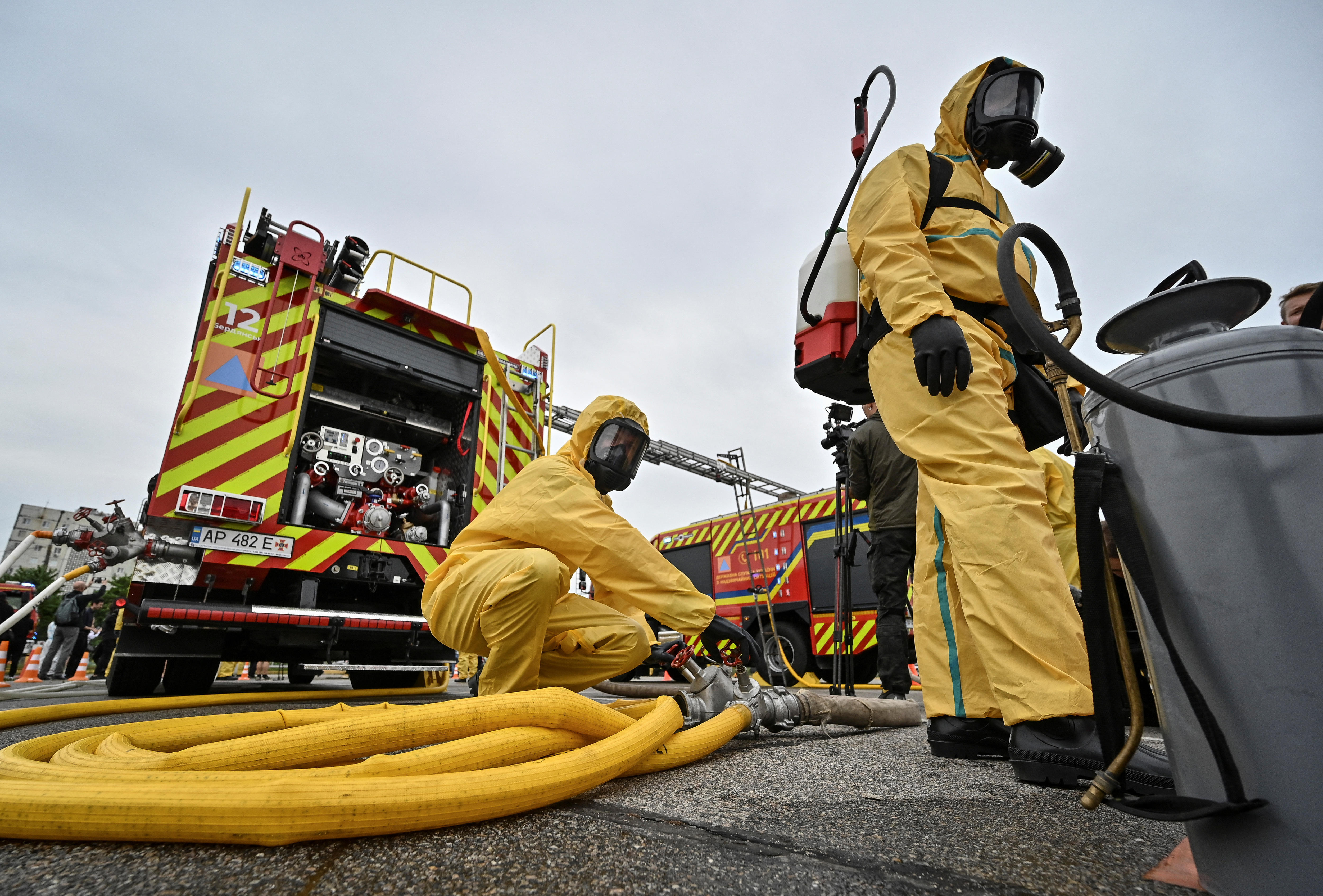 Two men in bright yellow hazmat suits attend to a large yellow firefighting hose in a car park.