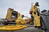 Two men in bright yellow hazmat suits attend to a large yellow firefighting hose in a car park.