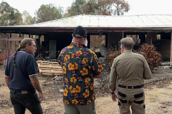 Three men stand outside burnt out structure.