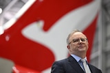 Anthony Albanese, wearing a suit, stands in front of a Qantas plane, with the logo visible. 