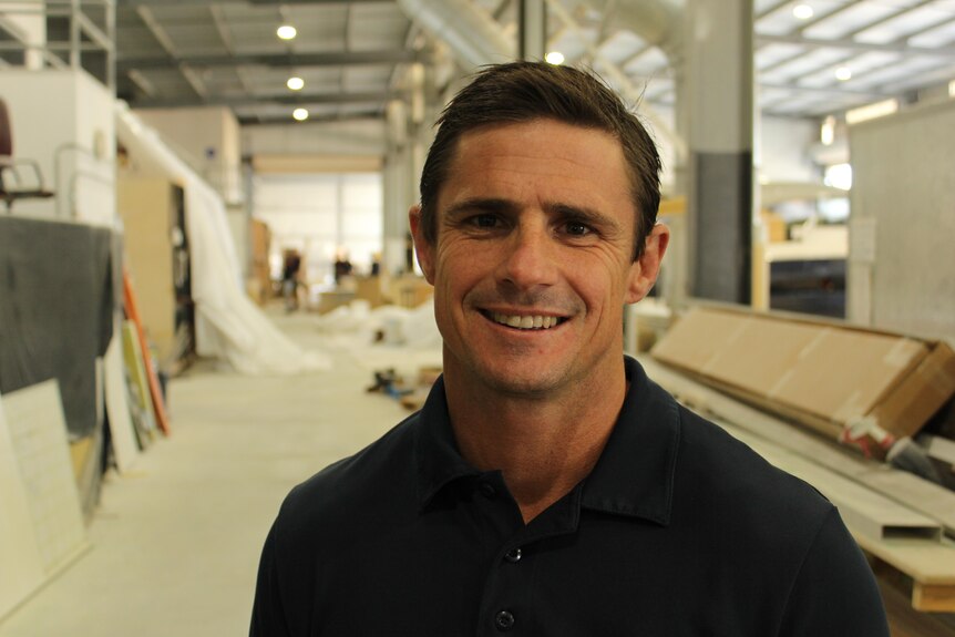 A man in a black shirt with brown hair and brown eyes smiling, with a boat-building workshop in the background
