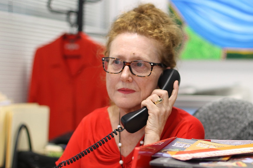 A woman with red hair and spectacles on sits at a desk in an office holding a phone receiver to her face..