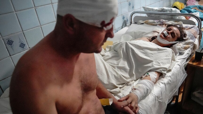 A shirtless man with his head bleeding through a bandage sits on a hospital bed in a corridor beside another victim.