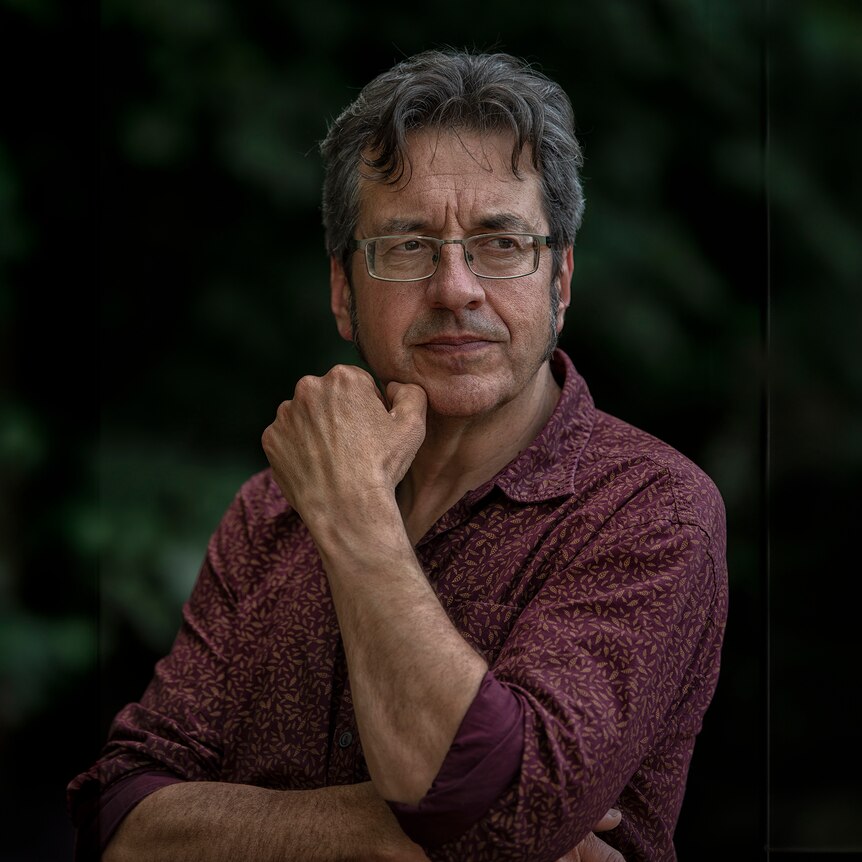 Grey haired man with glasses, maroon coloured shirt in profile, green leafy background, hands mimic the gesture of the thinker