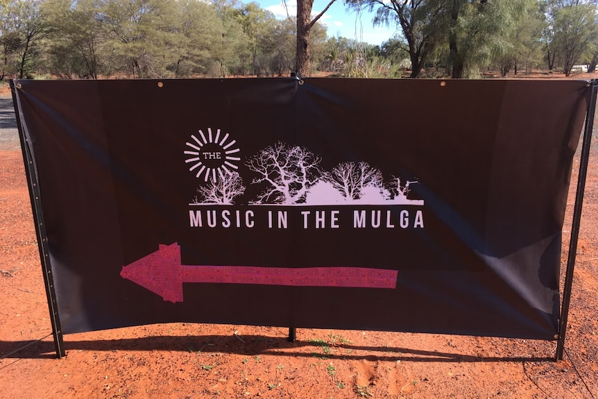 A black canvas sign that says 'Music in the Mulga' with a red arrow pointing left.