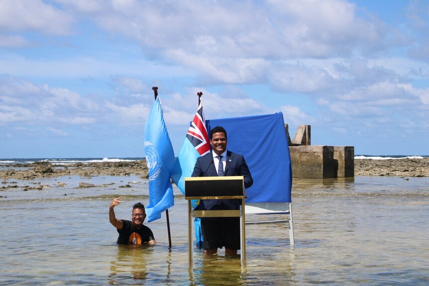 A man at a lectern and two flags gives a speech while standing knee-deep in the sea and another man to his right is chest-deep