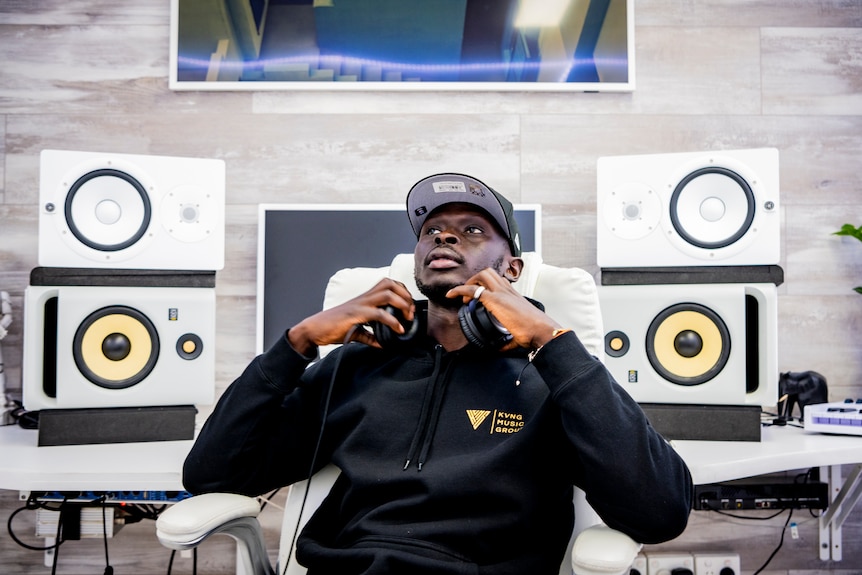 A man sits in a chair holding the headphones around his neck. Behind him is a computer and several large speakers.