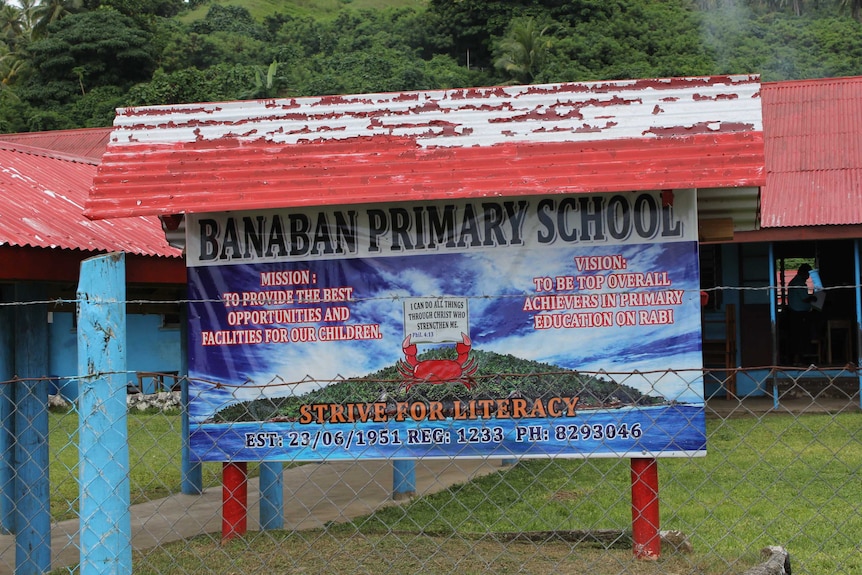 A sign that reads 'Banaban primary school', behind a gate, with trees and hills in the background.