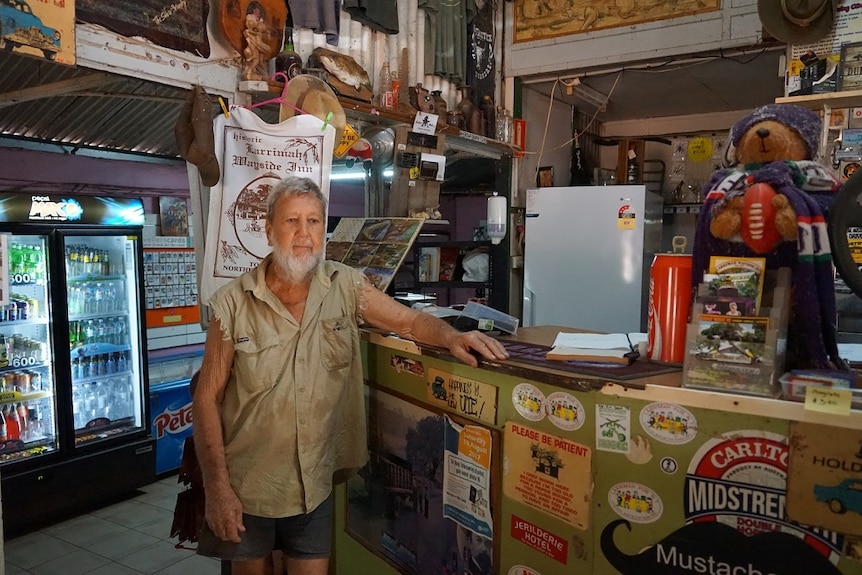 A man stands with his left arm reaching along the counter of a shop.