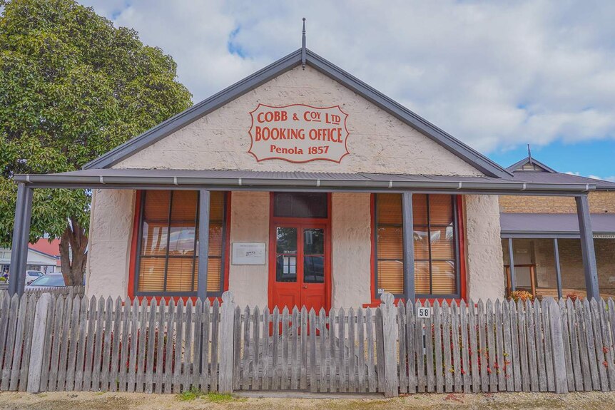 An old stone building with bright red window panes and wooden doors with a sign reading 'Cobb and Co Booking Office Penola 1857'