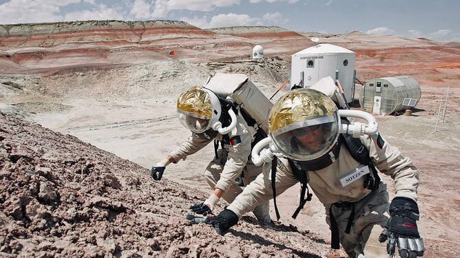 Two men in spacesuits scrabble up a pile of rocks in the desert