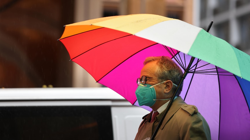 A man in a face mask walks down the street in the rain under a rainbow umbrella