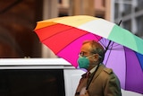 A man in a face mask walks down the street in the rain under a rainbow umbrella