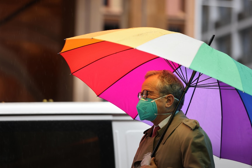 A man in a face mask walks down the street in the rain under a rainbow umbrella