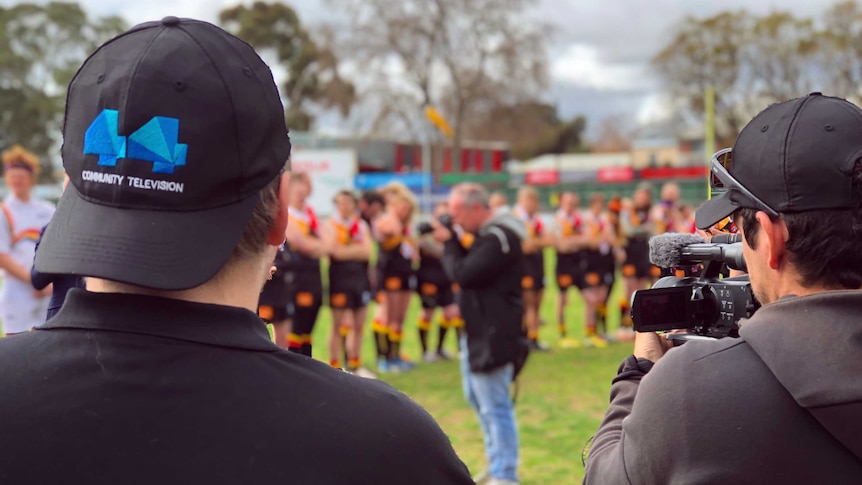 A man with a backwards 44 community television hat and a camera operator film a sports team on an oval.