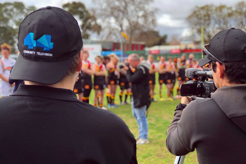 A man with a backwards 44 community television hat and a camera operator film a sports team on an oval.