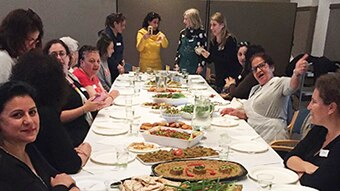 A group of smiling women sit at a long table about to eat a banquet of food.