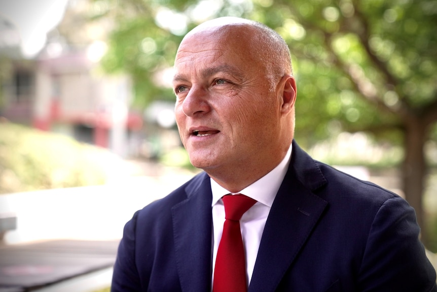 A man in suit wearing a red tie and speaking outdoor. 