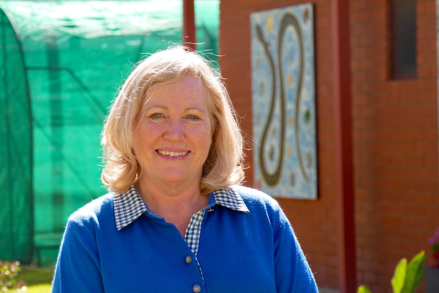 A blonde woman wearing a blue sweater in yard with an aboriginal artwork in the background. 