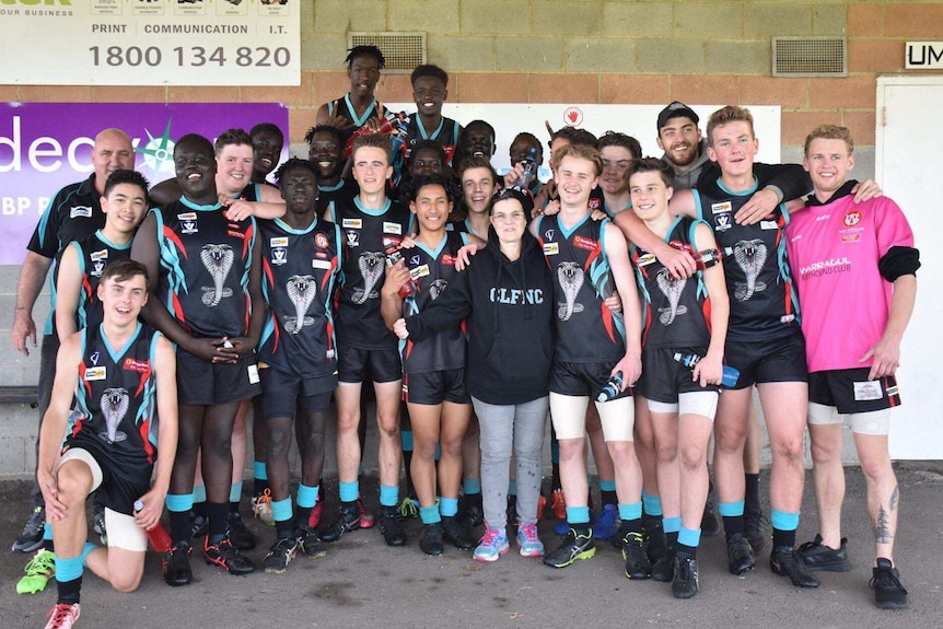 The under-16s team and support staff stand together in their uniforms for a group photo in front of their clubrooms.