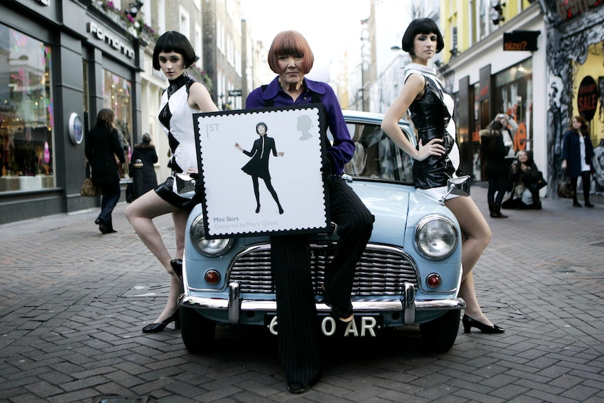Three women with bobs in front of a car, the one in the centre holding a poster 