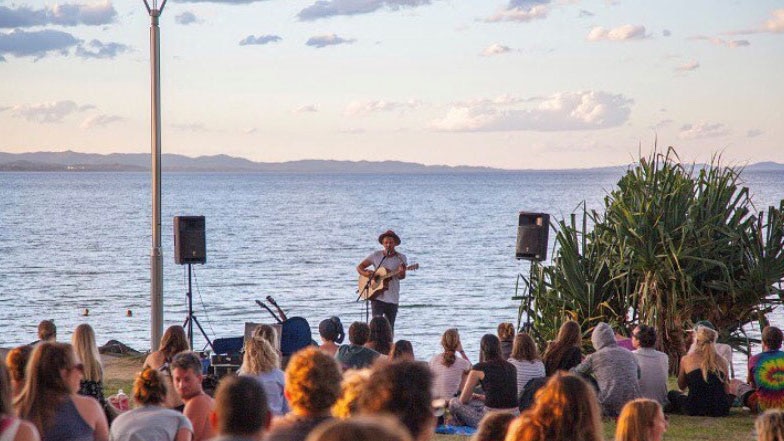 A crowd sit on a lawn by the sea watching a busker standing at the shore play guitar.
