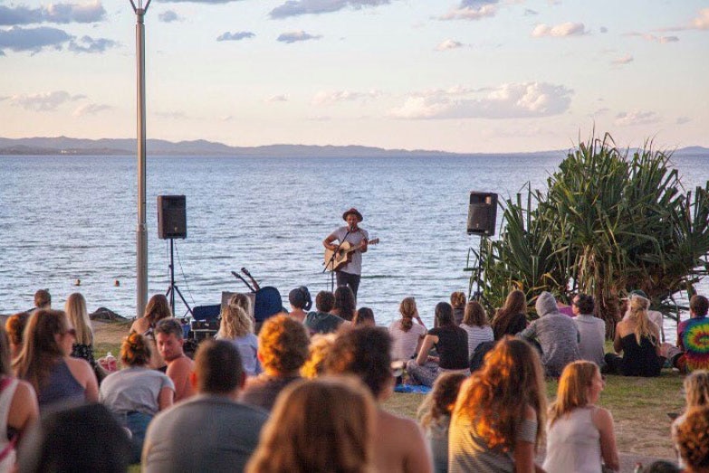 A crowd sit on a lawn by the sea watching a busker standing at the shore play guitar.