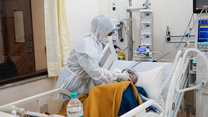 An old man in a hospital bed, with a health worker in full PPE at his side