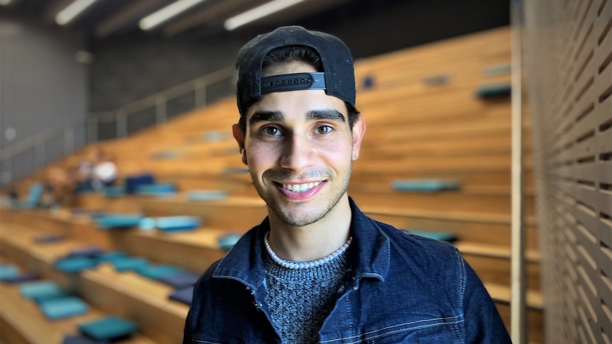 A portrait of Isaiah smiling, wearing a denim jacket, a cap and a string of pearls, standing in front of an assembly hall. 