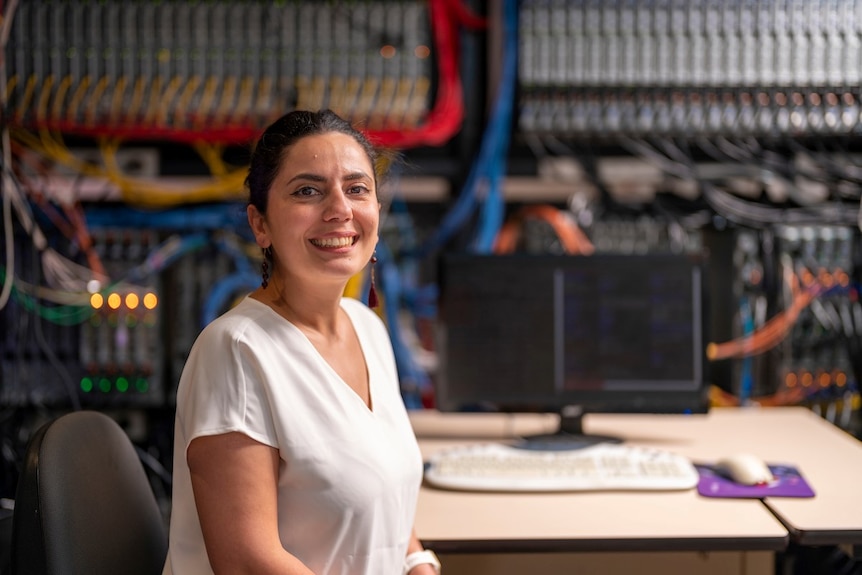 A woman in a white t-shirt sitting at a desk and smiling at the camera