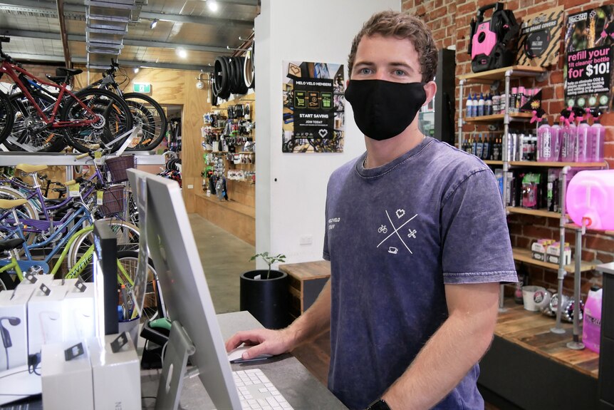 A masked man standing behind a computer with bikes and equipment in the background.