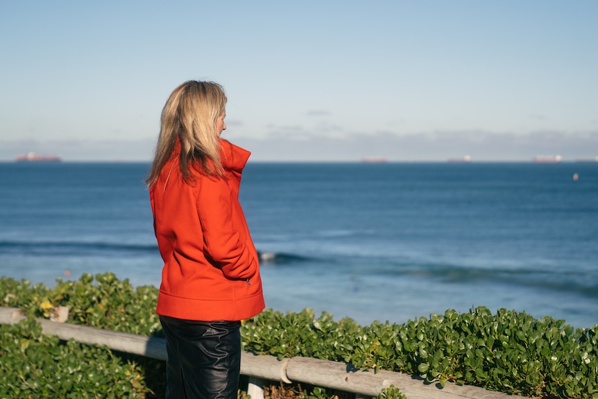 Bourby wears a bright red jacket and black pants, facing away from the camera, standing on the coast looking into water.