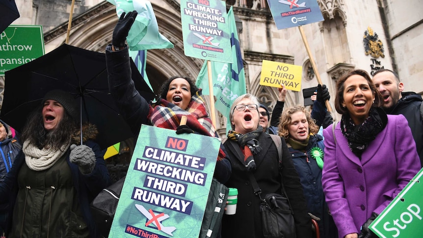 People holding signs saying "no climate-wrecking third runway" cheer outside a courthouse.