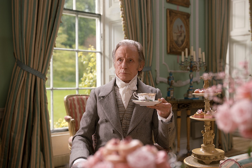 An older man in three piece grey Regency England period costume sits in period interior and holds teacup on saucer.