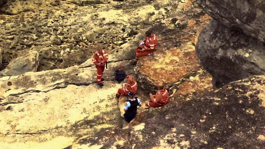 SES personnel and a police officer stand and sit on coastal rocks.