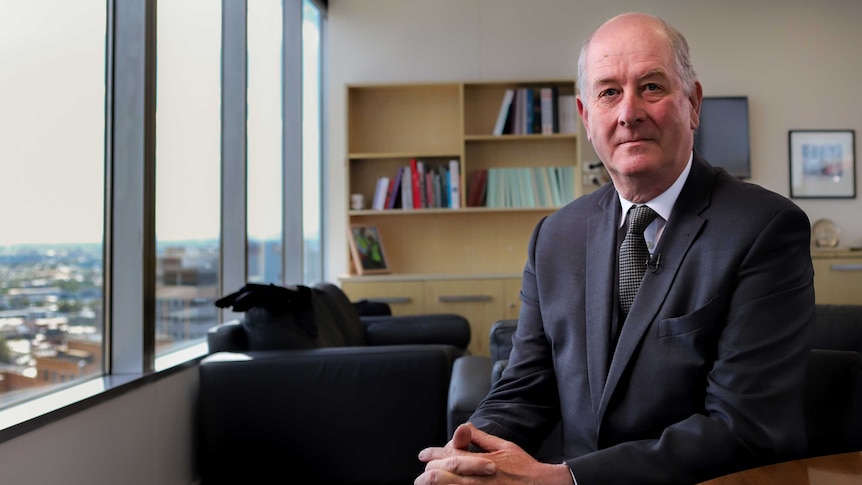 Richard Wynne MP, wearing a suit and tie, sits in an office with windows looking out over a city view.